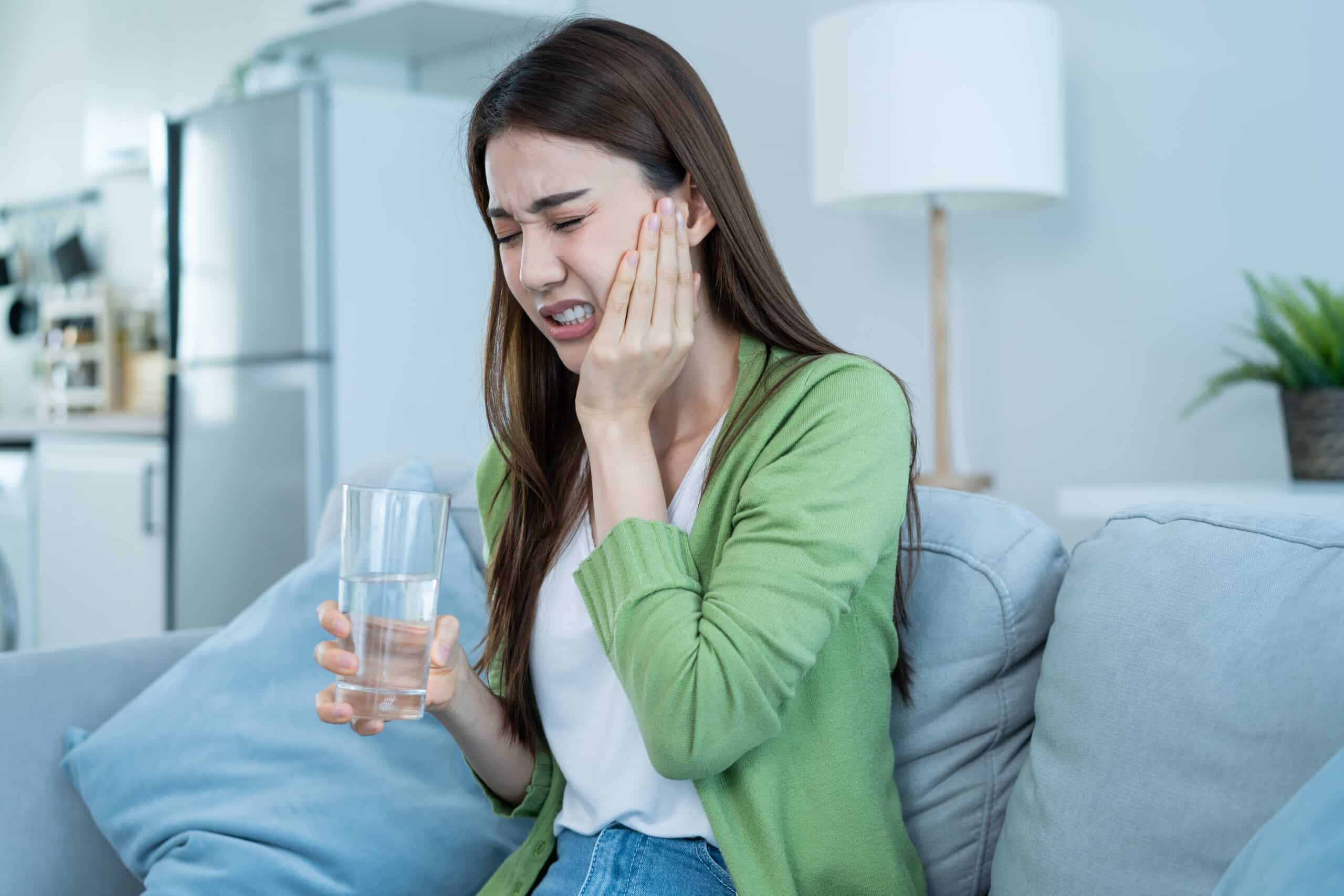 Woman holding her cheek in pain while sitting on a sofa, experiencing a toothache and drinking water at home.