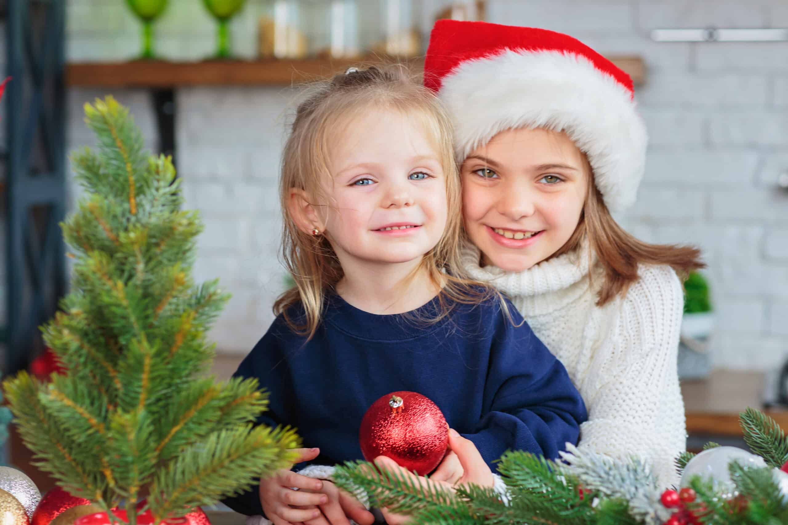 Two smiling children wearing winter clothes and a Santa hat, posing together beside a small Christmas tree with holiday decorations indoors.