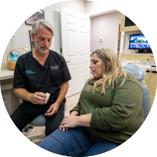 Male dentist in scrubs consulting with female patient in dental office treatment room.