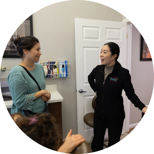 Two women conversing in a dental office hallway, one wearing a black jacket with dental studio branding.