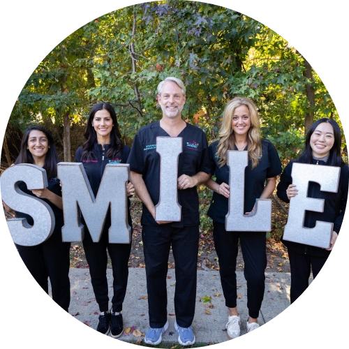 Five dental team members holding large letters spelling "SMILE" outdoors against green foliage background.