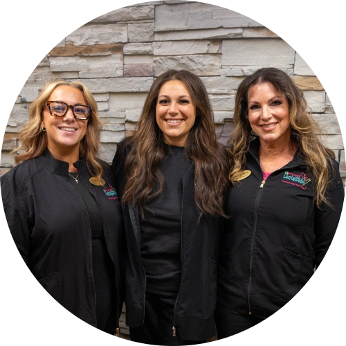 Three female dental professionals smiling at camera against stone wall backdrop.
