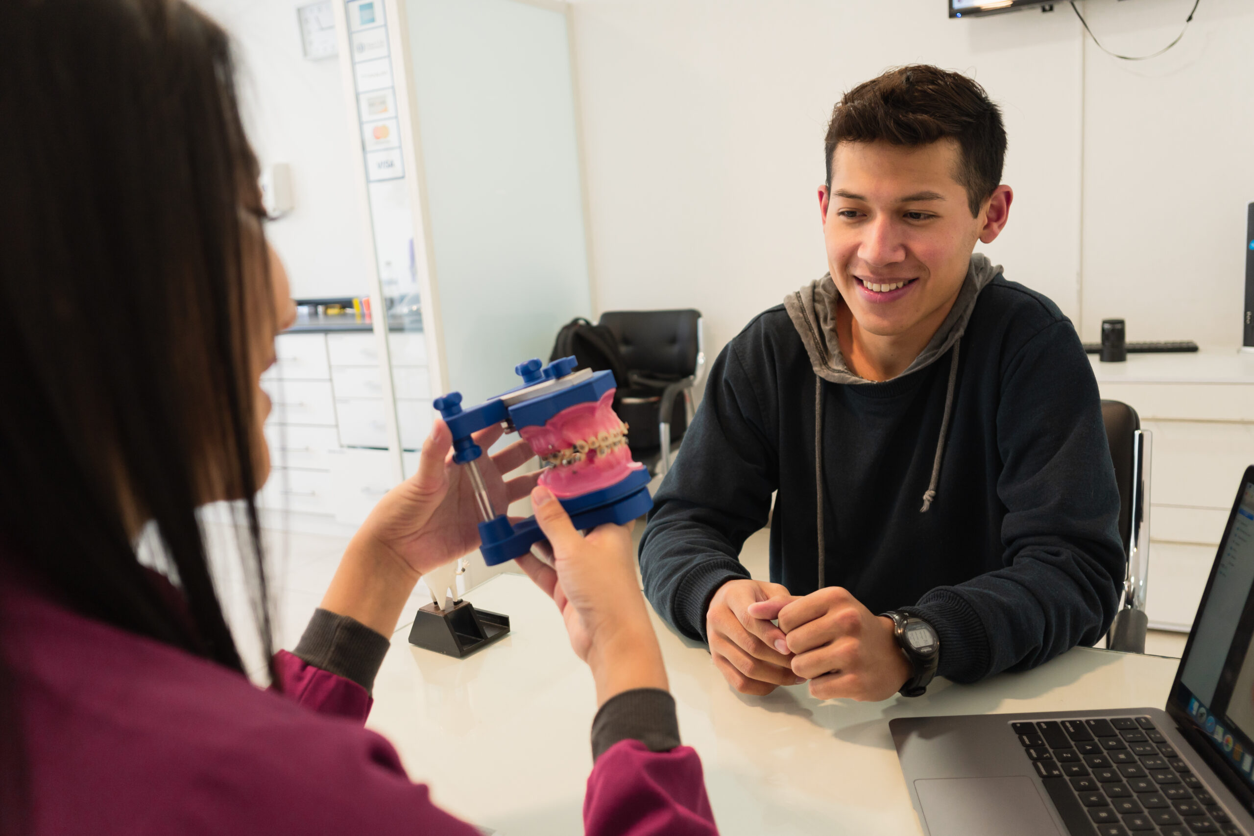Adult patient smiling during orthodontic consultation while dentist explains braces using a dental model in a modern office setting.