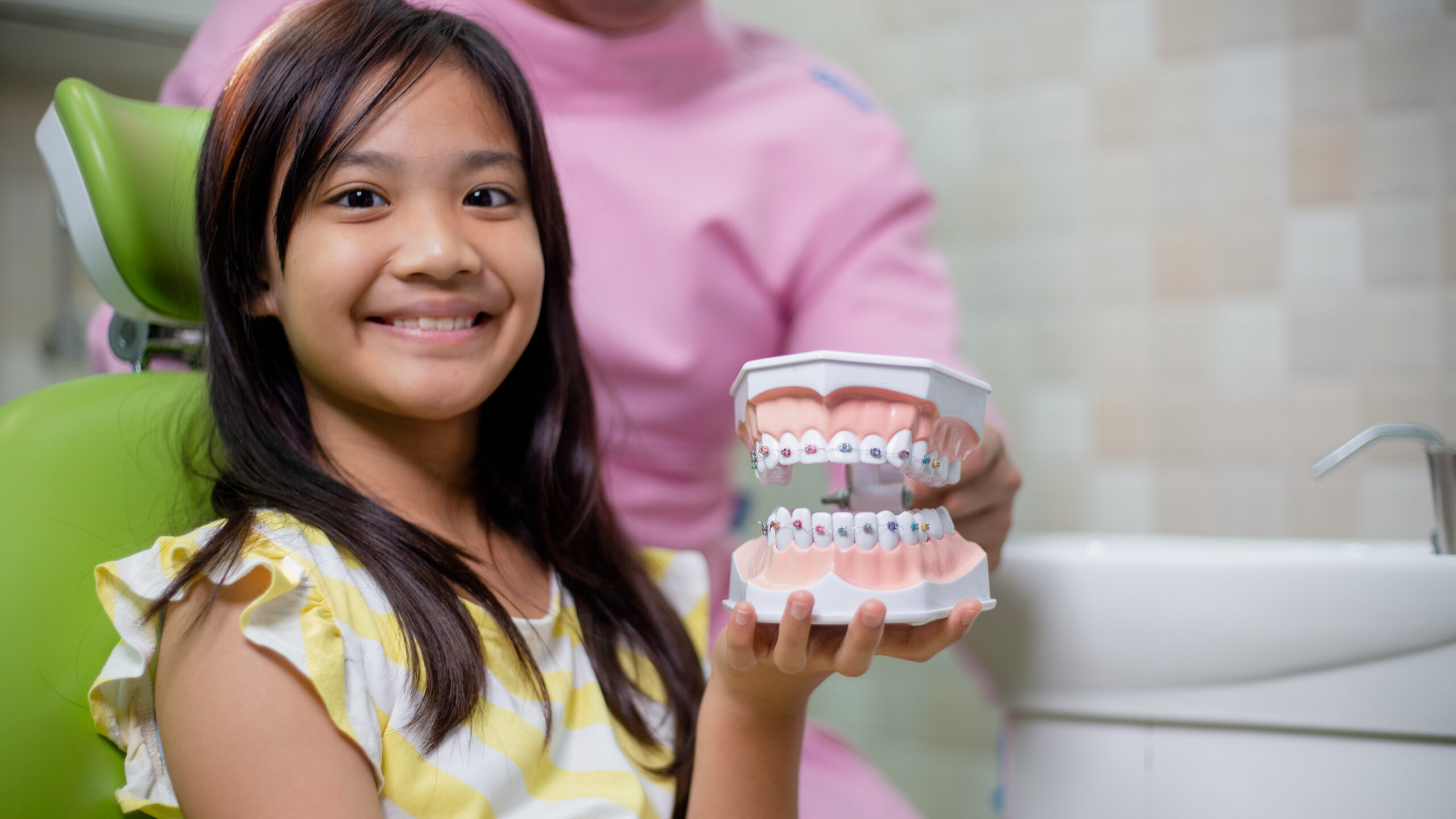 Child smiling in a dental chair while holding a model showing braces during an orthodontic checkup.