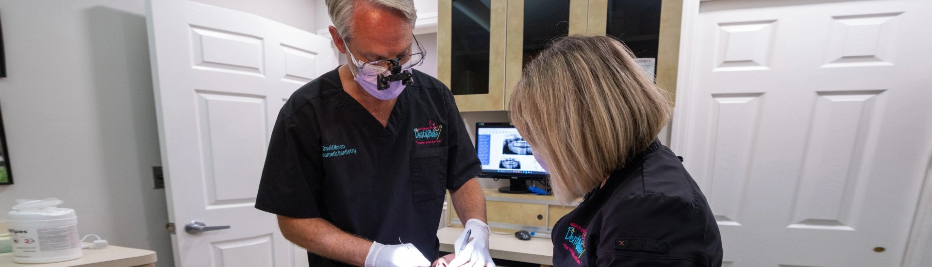 Two dental professionals wearing scrubs and protective gear working together to treat a patient