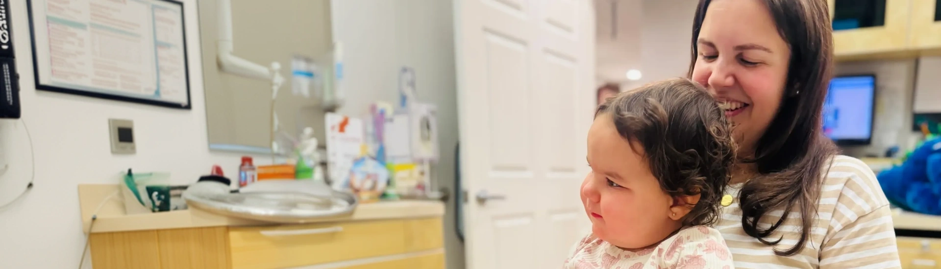 A smiling mother holding her young toddler inside a dental exam room