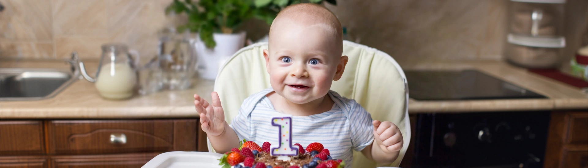 A cheerful one-year-old infant sitting behind a small birthday cake in a kitchen setting
