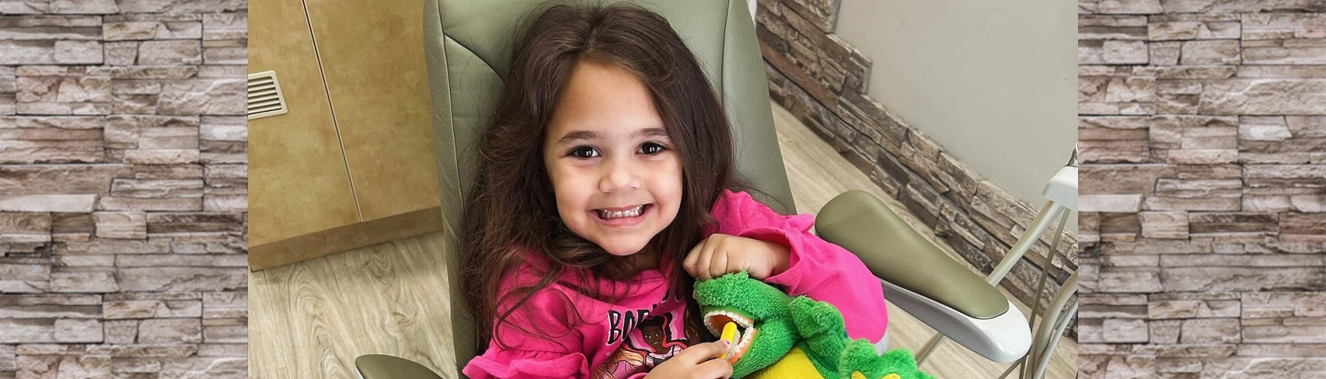 A happy, smiling young girl with a plush toy sitting comfortably in a pediatric dental chair