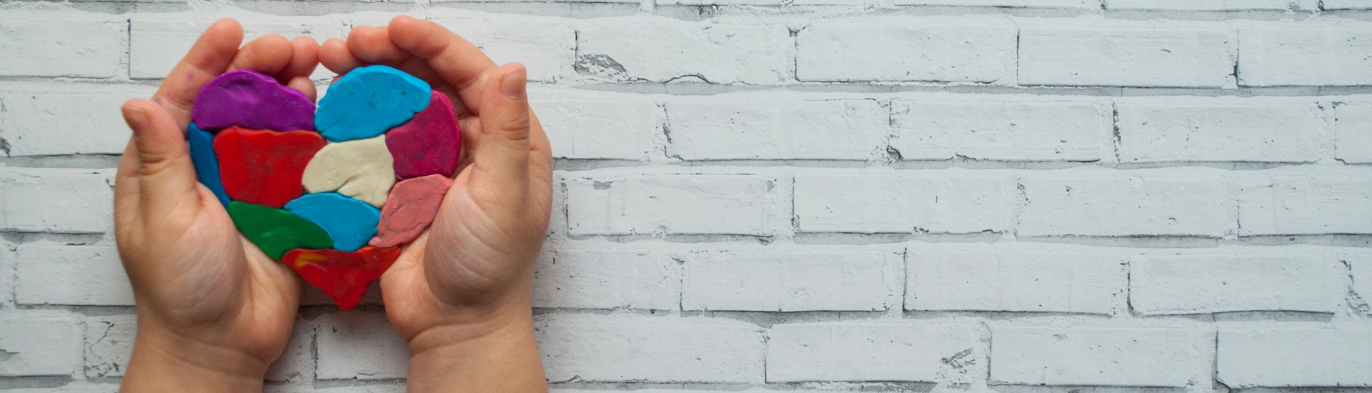 Two hands cupping a colorful puzzle-piece styled heart against a white brick backdrop