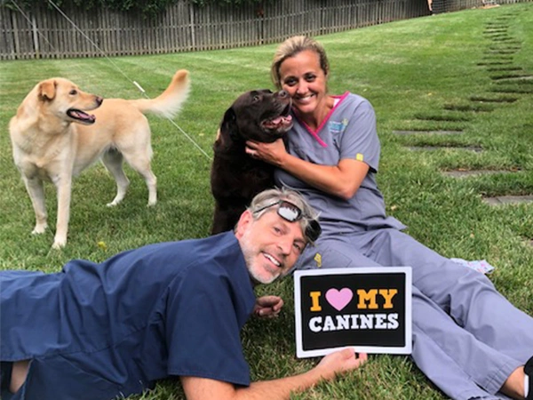 Dental team members posing with their dogs outdoors while holding an "I Love My Canines" sign.